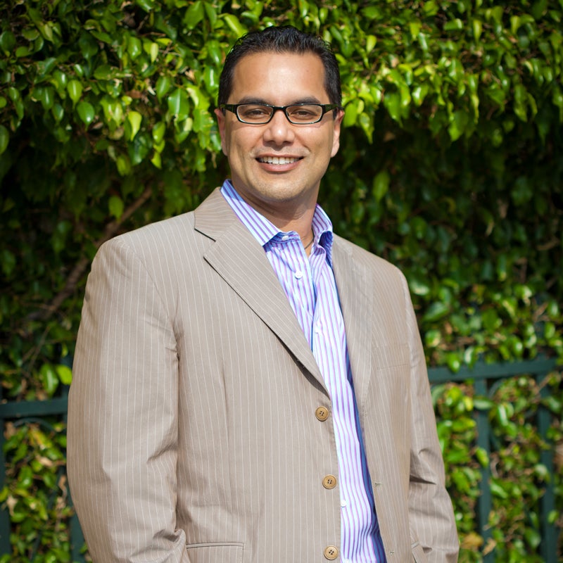 A suited Dr. Sharma with glasses poses in front of vibrant green bushes in Jupiter, FL.