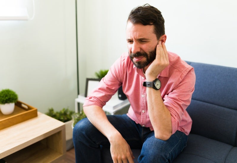 A man sitting on a couch, holding his ear, highlighting tinnitus treatment options in Jupiter, FL.